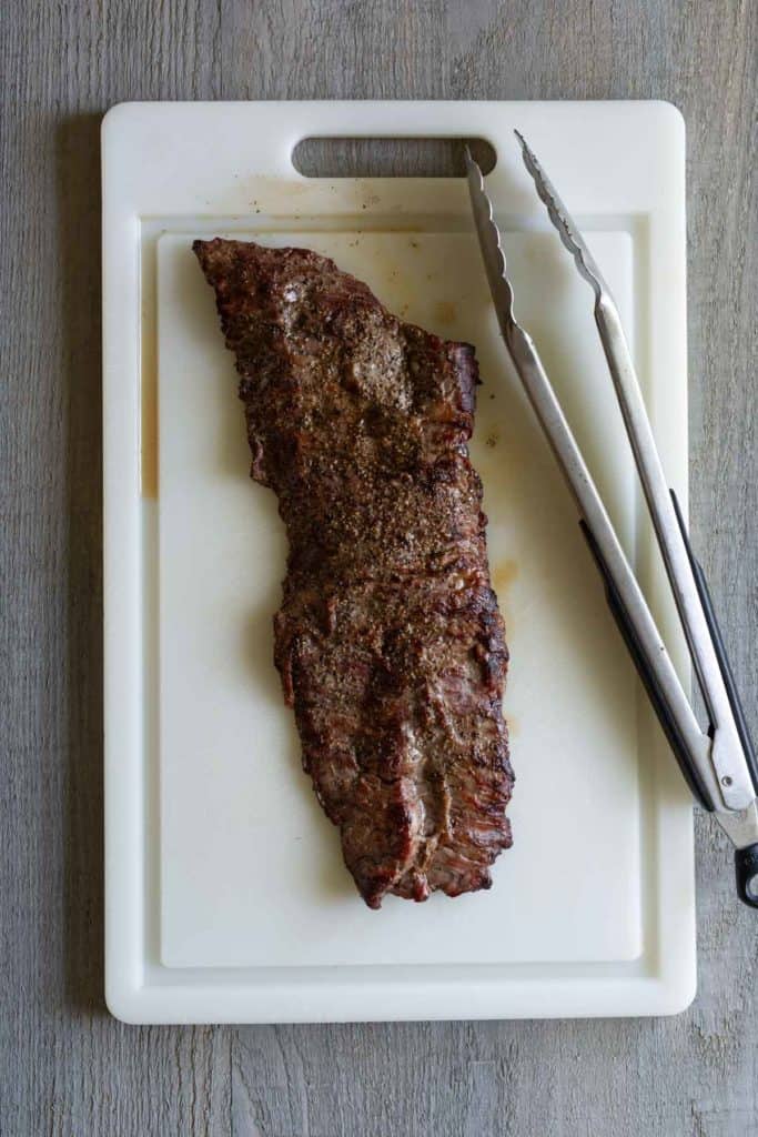 A piece of cooked meat with seasoning is placed on a white cutting board next to a pair of metal tongs.