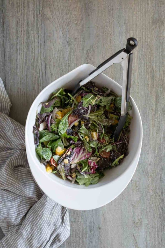 A white bowl filled with a mixed green salad, including leafy greens, red onions, corn, and grilled vegetables. Black tongs rest in the bowl, and a striped cloth napkin is next to it on a wooden surface.