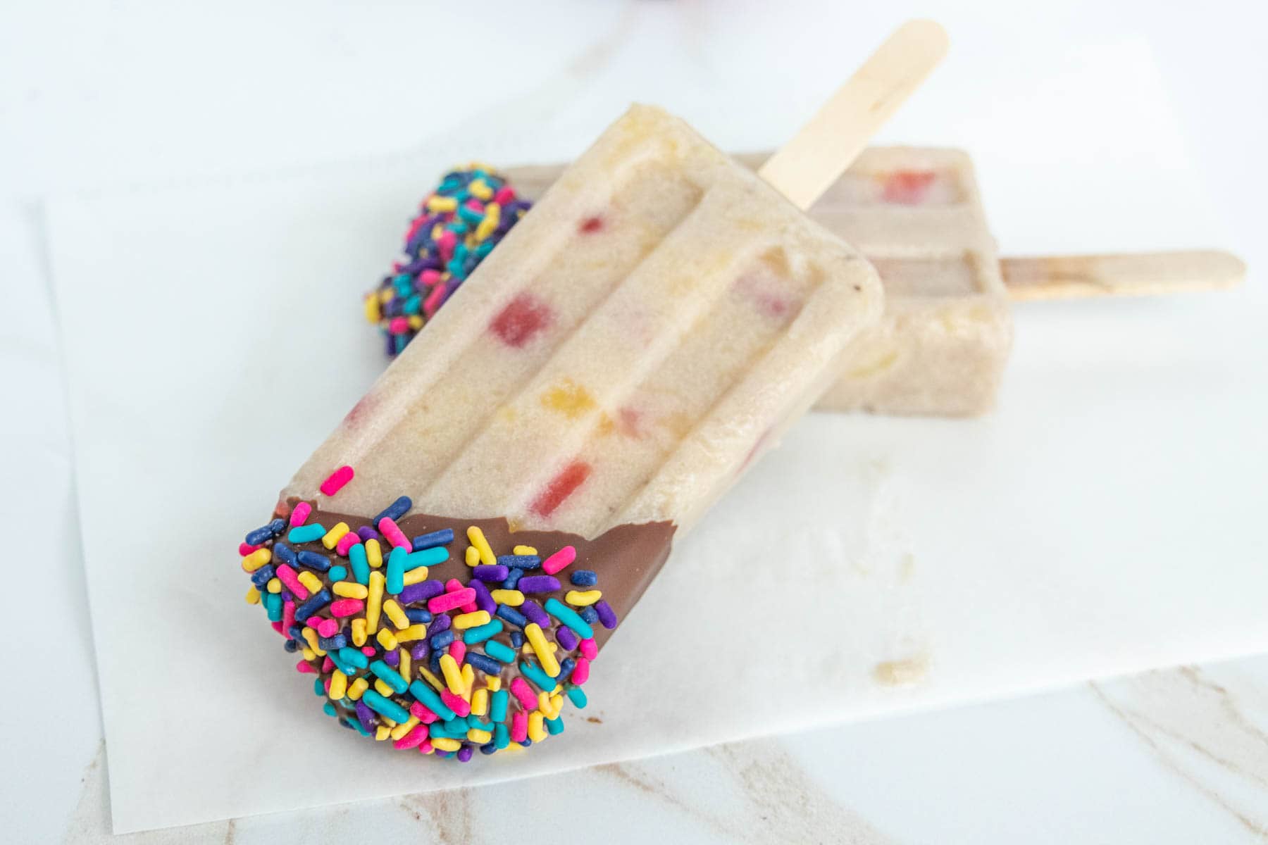 Two popsicles with pieces of fruit, one dipped in chocolate and colorful sprinkles, placed on a white surface.