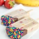 Two ice cream bars dipped in chocolate and sprinkled with colorful candy sprinkles, with strawberries and a banana in the background.