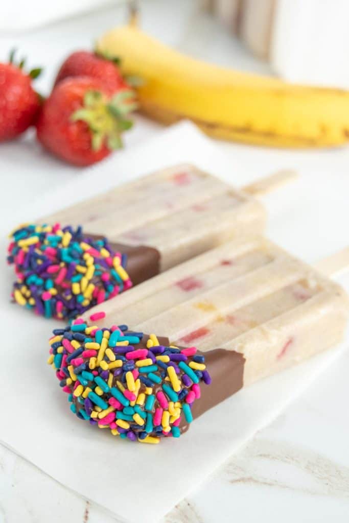 Two ice cream bars dipped in chocolate and sprinkled with colorful candy sprinkles, with strawberries and a banana in the background.