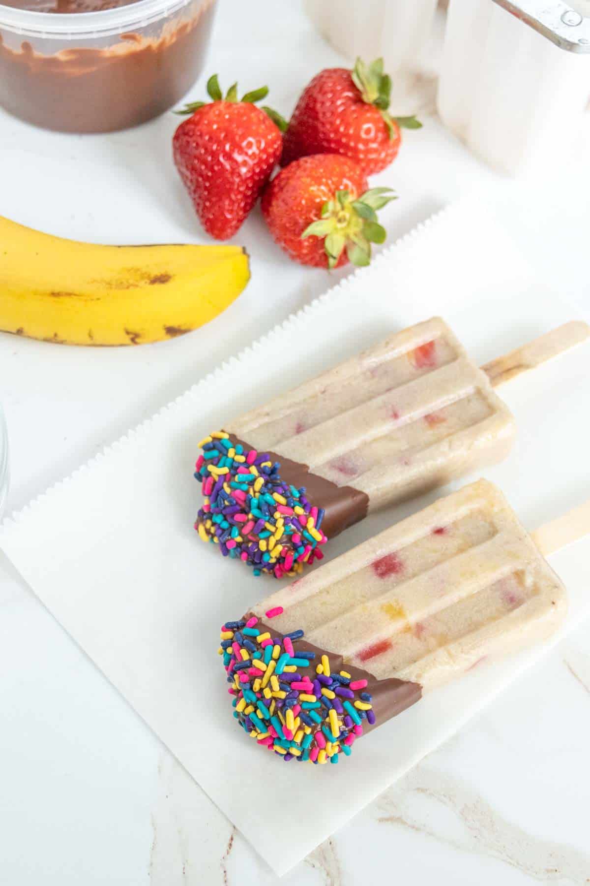 Two popsicles partially dipped in chocolate and colorful sprinkles are placed on a piece of parchment paper beside a banana and fresh strawberries.