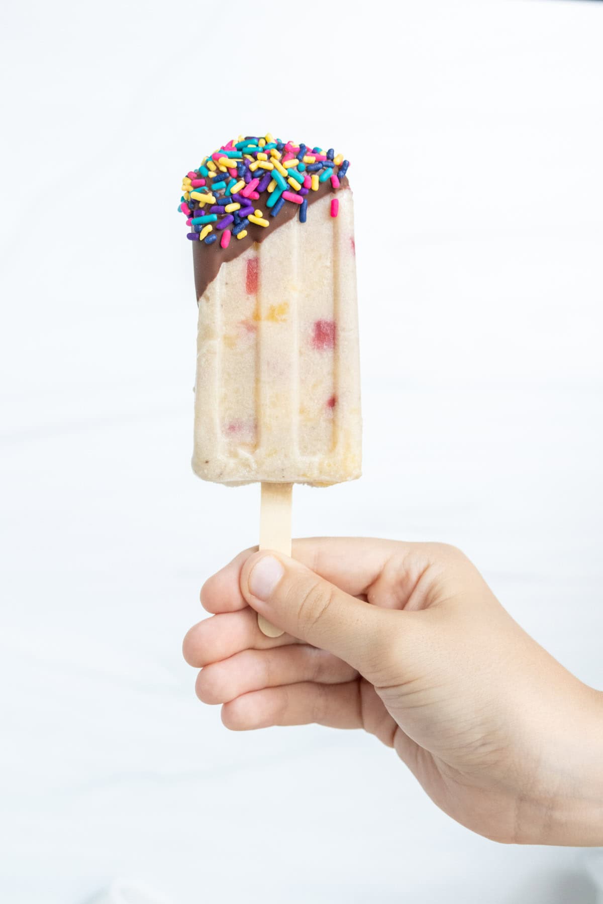 A hand holding a popsicle partially dipped in chocolate and covered with colorful sprinkles.