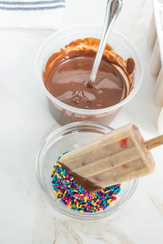 A popsicle being dipped in a small bowl of multicolored sprinkles next to a container of melted chocolate with a spoon.