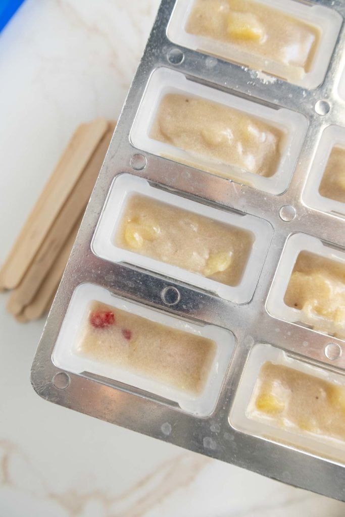 Close-up of a metal ice cream mold tray filled with popsicle mixtures, featuring visible fruit pieces, positioned on a white table. Wooden sticks are placed beside the tray.