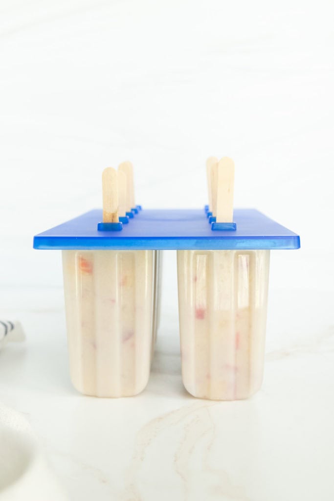 A popsicle mold with a blue lid, containing partially frozen popsicles with wooden sticks, is set on a marble surface.