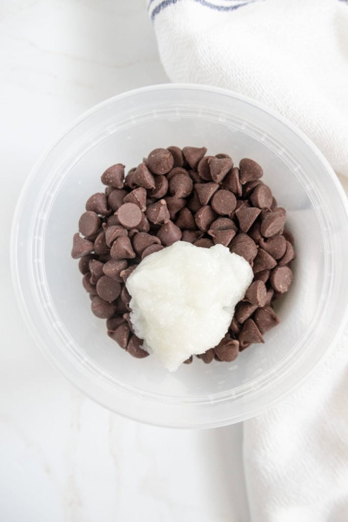 A plastic container holds chocolate chips with a dollop of coconut oil on top, placed on a white surface next to a white cloth.