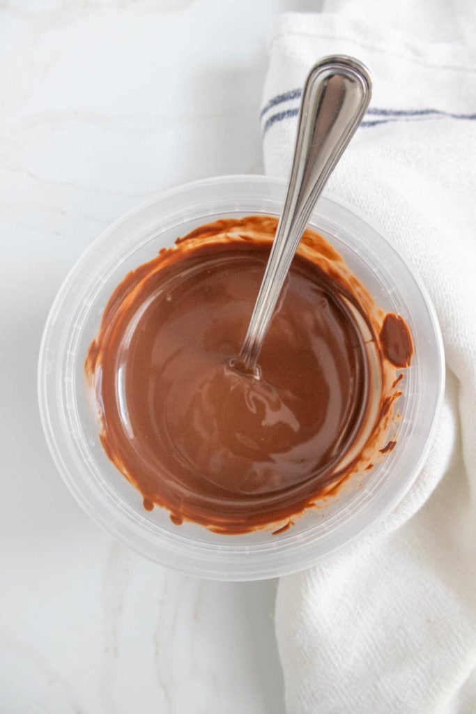 A plastic container filled with melted chocolate and a spoon, placed next to a white cloth.