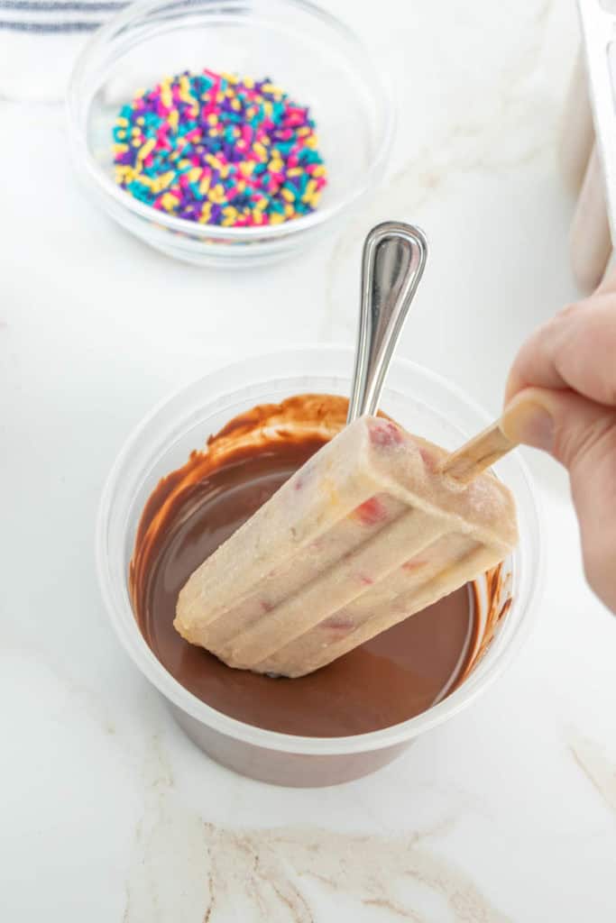 A hand dips a popsicle with visible colorful bits into a container of melted chocolate. A bowl of multicolored sprinkles is in the background.