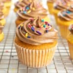 A cupcake with chocolate frosting and colorful sprinkles is on a cooling rack, surrounded by similar cupcakes in the background.
