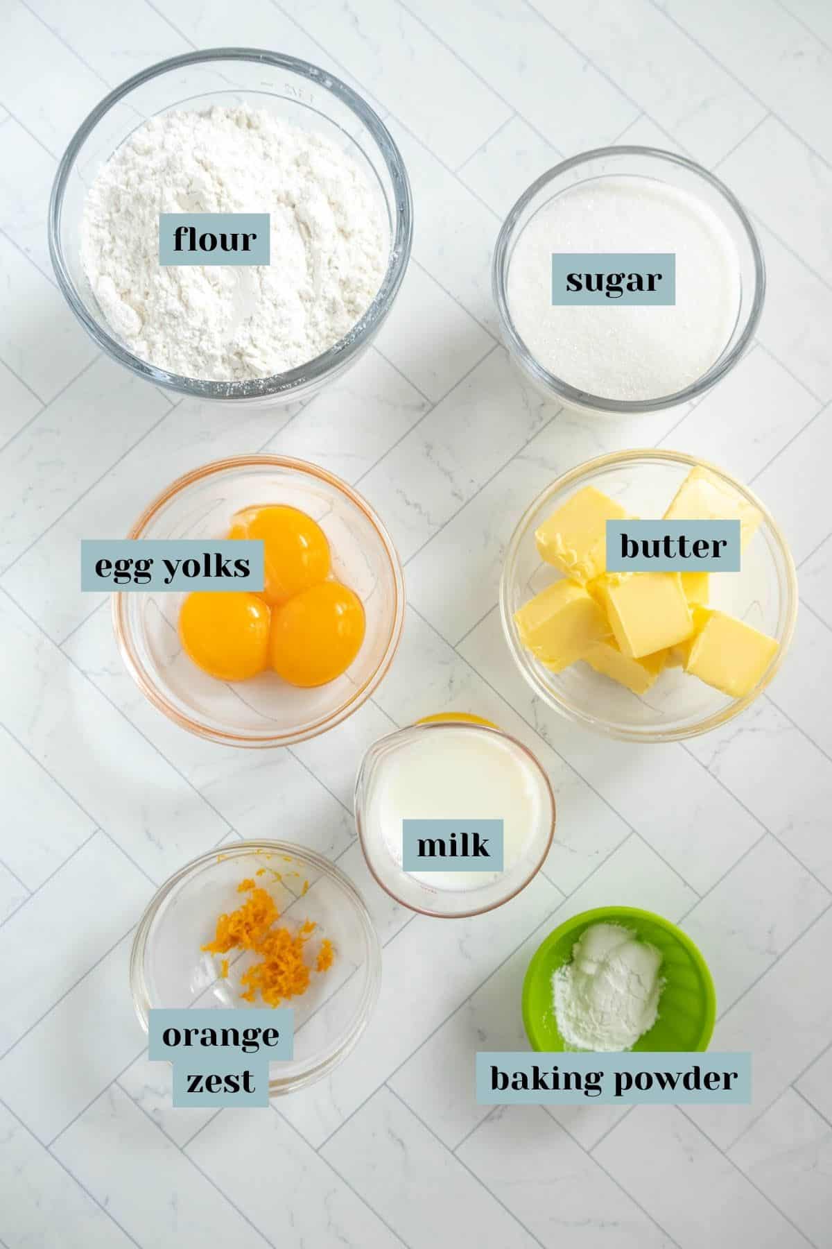 A flat lay of baking ingredients in bowls on a white surface, including flour, sugar, egg yolks, butter, milk, baking powder, and orange zest.