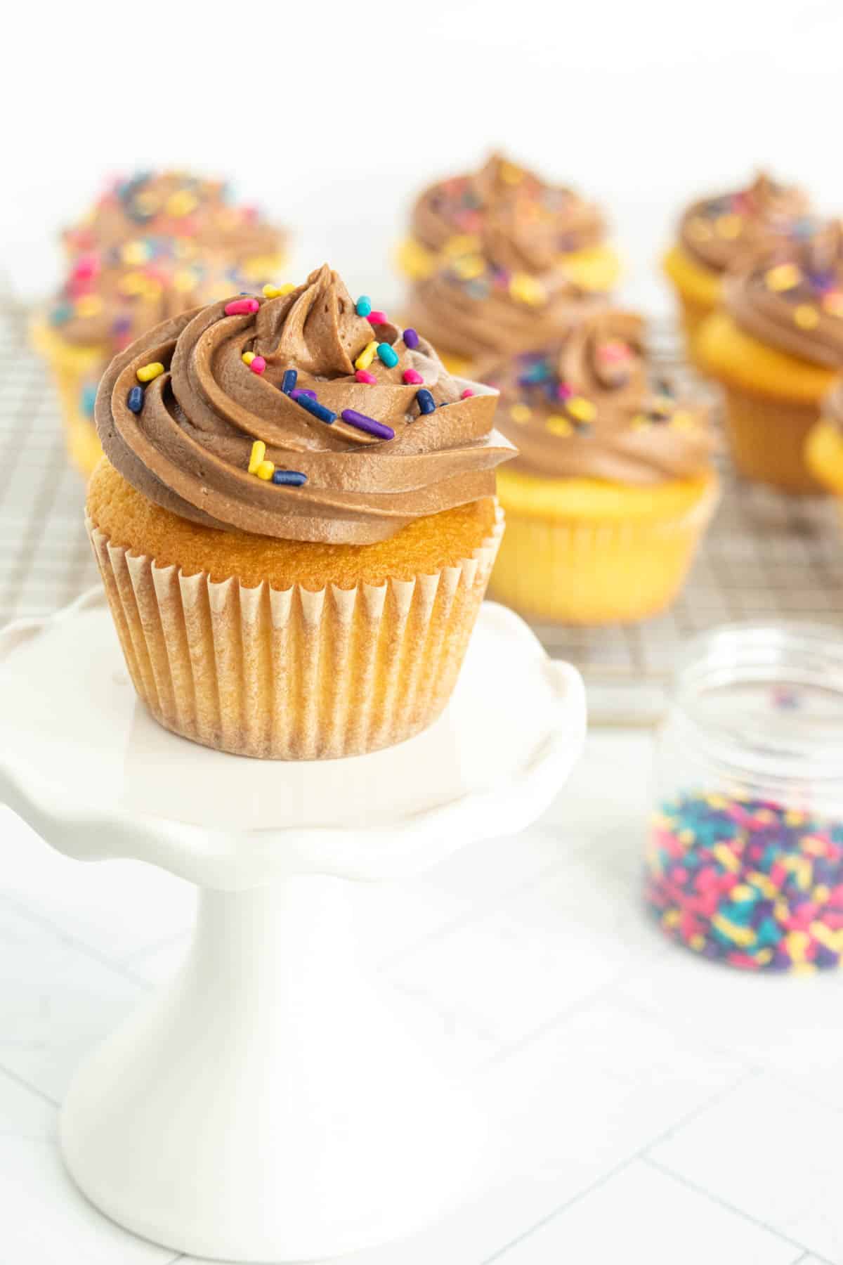 A vanilla cupcake with chocolate frosting and colorful sprinkles on a white stand, with more cupcakes and a jar of sprinkles in the background.