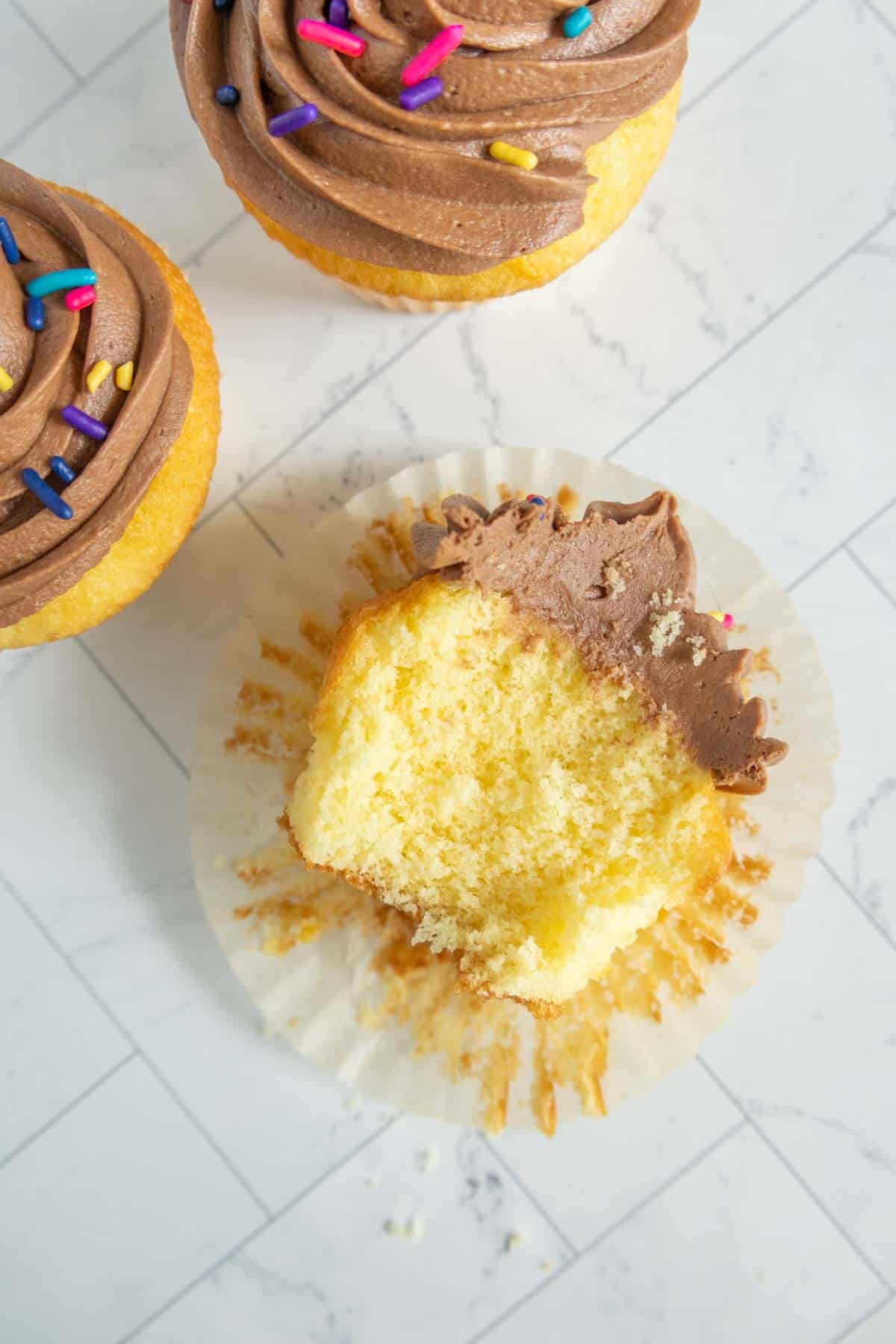A partially eaten yellow cupcake with chocolate frosting and sprinkles, placed on a white wrapper, with two additional frosted cupcakes nearby on a light marble surface.