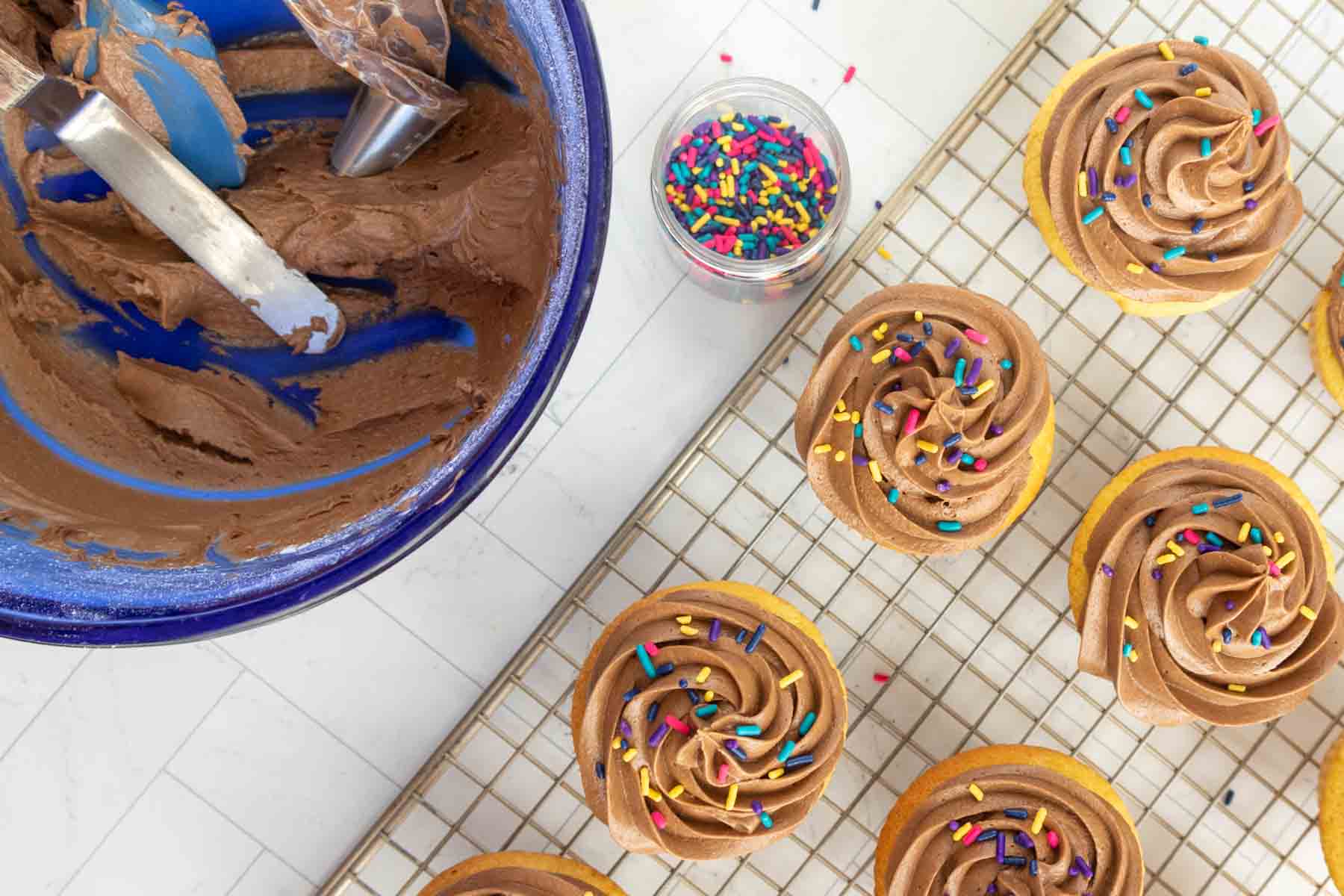Cupcakes topped with chocolate frosting and colorful sprinkles rest on a cooling rack next to a bowl of leftover frosting and a small container of sprinkles.