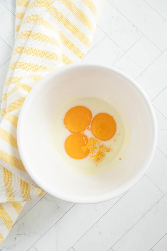 A white bowl with three cracked eggs on a tiled surface, next to a yellow and white striped kitchen towel.