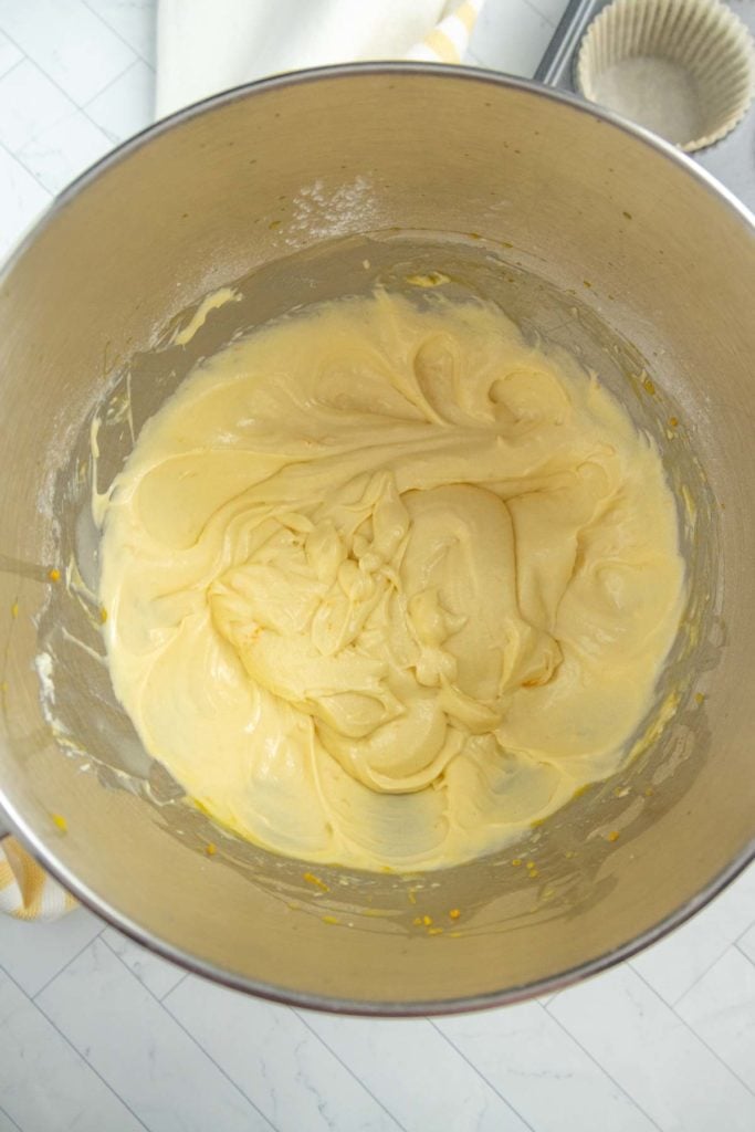 A stainless steel mixing bowl contains a creamy yellow batter. The bowl is placed on a white surface, with baking supplies visible in the background.