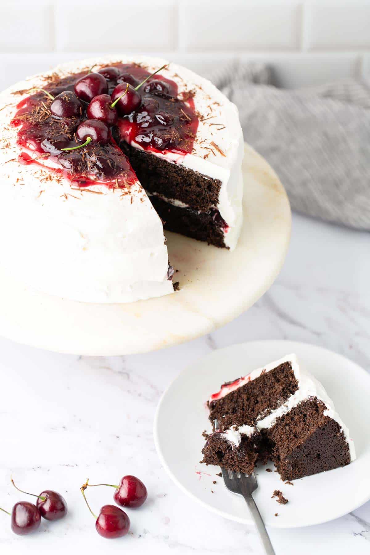Chocolate cake with cherry topping and white frosting on a marble stand, with a slice cut out and served on a plate next to it. Fresh cherries are placed on and beside the cake.