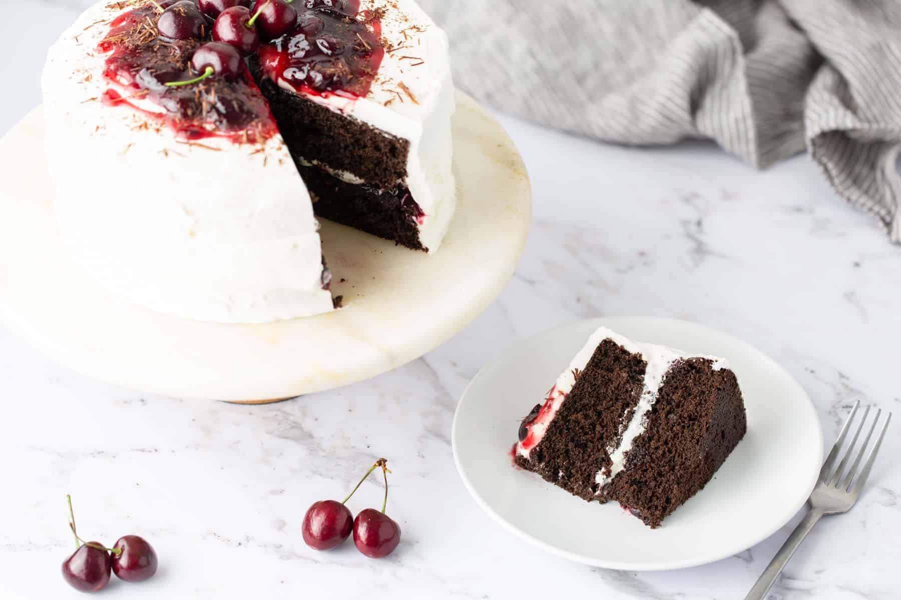 A chocolate cake with white frosting and cherries on top sits on a marble stand with a slice cut out and placed on a plate beside it. A few cherries are scattered on the marble surface.