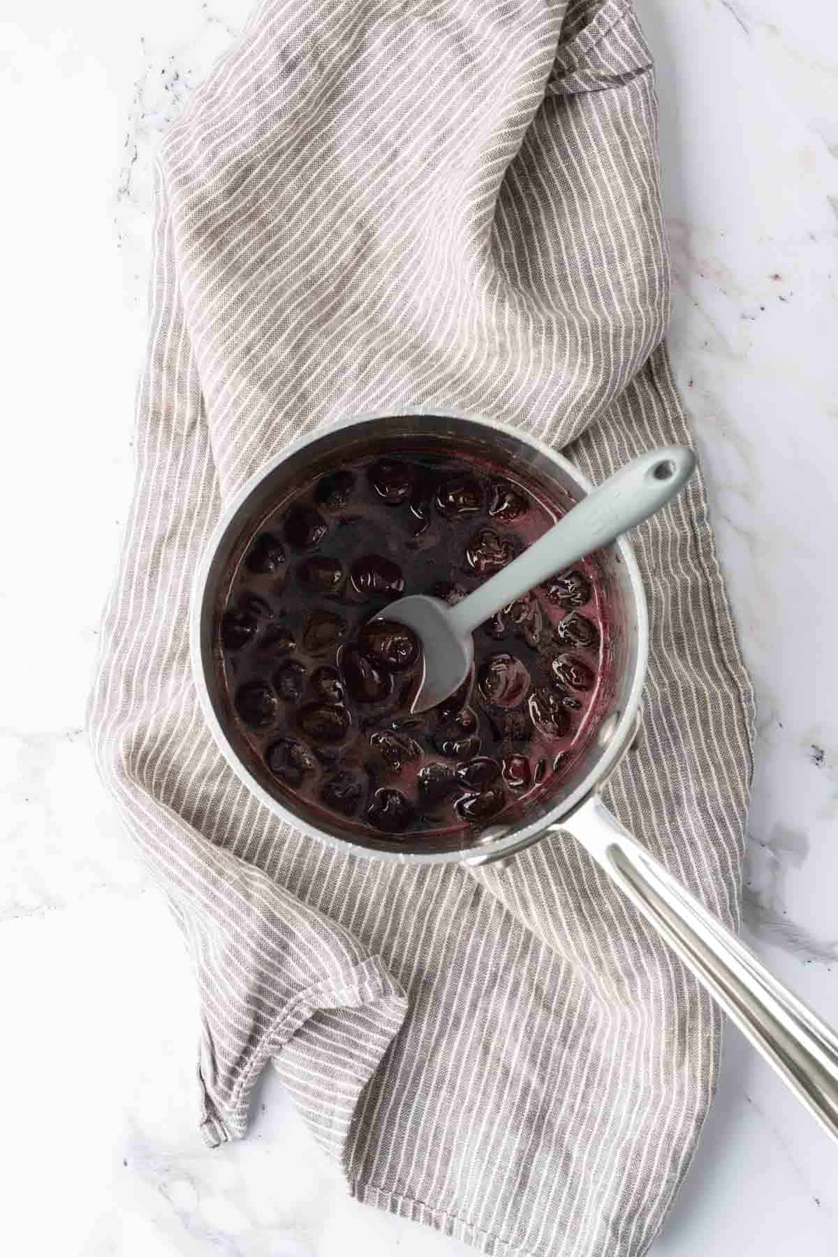 A saucepan filled with dark cherries in liquid, placed on a gray and white striped cloth. A silver spoon is in the saucepan.