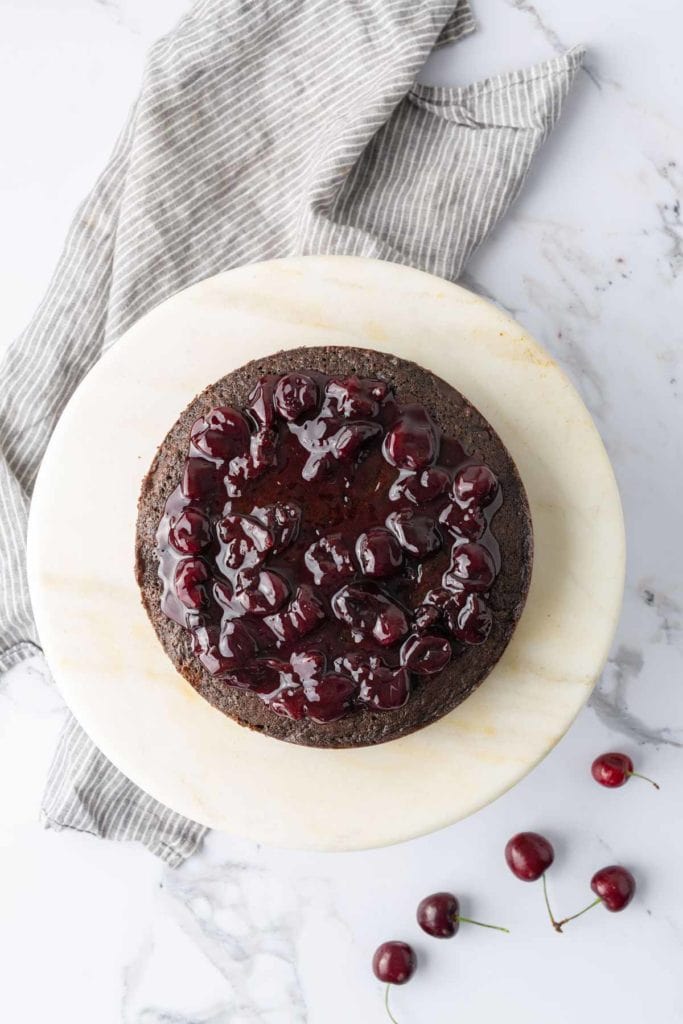 A round chocolate cake topped with cherries and a glaze, placed on a marble surface with a striped cloth in the background and a few cherries scattered nearby.