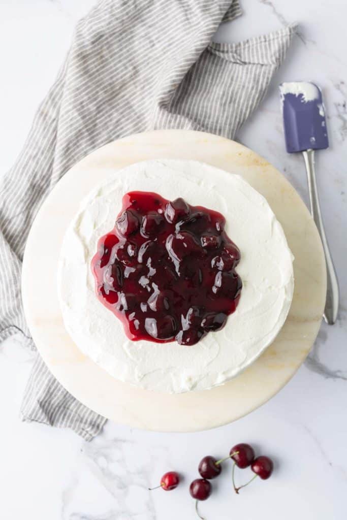 A round cake with white frosting topped with cherry filling is on a marble surface. A gray striped cloth and a spatula are beside the cake. Three cherries are on the surface near the cake.