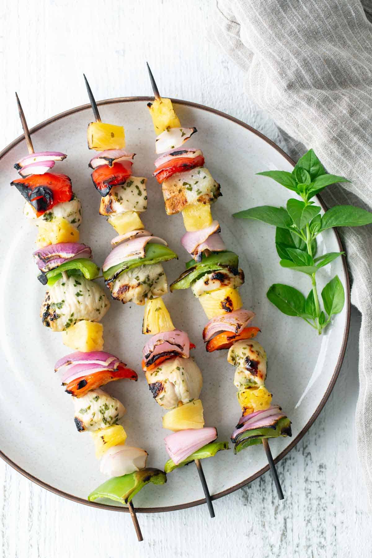 Three grilled chicken and vegetable kebabs on a white plate with a sprig of fresh basil and a striped cloth in the background.