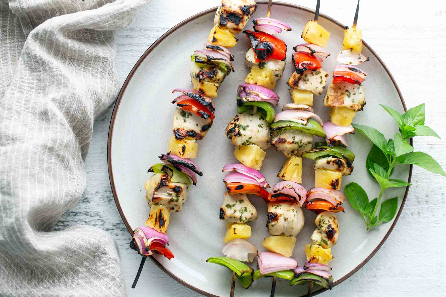 Plate with grilled chicken kebabs, skewered with bell peppers, red onions, and pineapple chunks, garnished with fresh herbs. A striped cloth napkin is beside the plate.