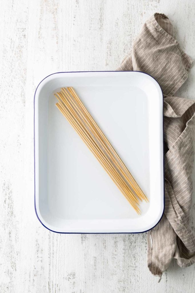 A handful of uncooked spaghetti lies in a white rectangular dish on a white wooden surface with a brown cloth to the right.