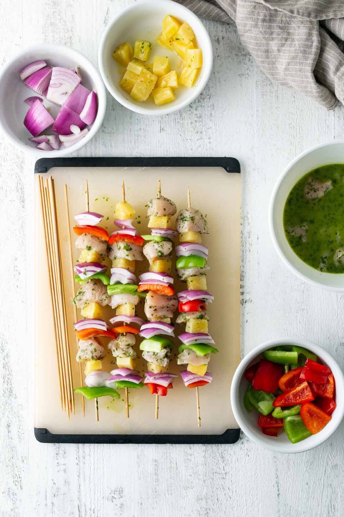 Four uncooked chicken kabobs with onions, peppers, and pineapples are arranged on a cutting board. Surrounding bowls contain additional sliced onions, diced pineapples, bell peppers, and marinade.