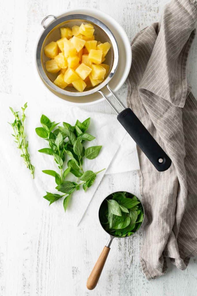 Overhead view of cubed pineapples in a strainer, fresh basil leaves on the table, and more basil in a measuring cup, all on a white textured surface with a gray cloth nearby.