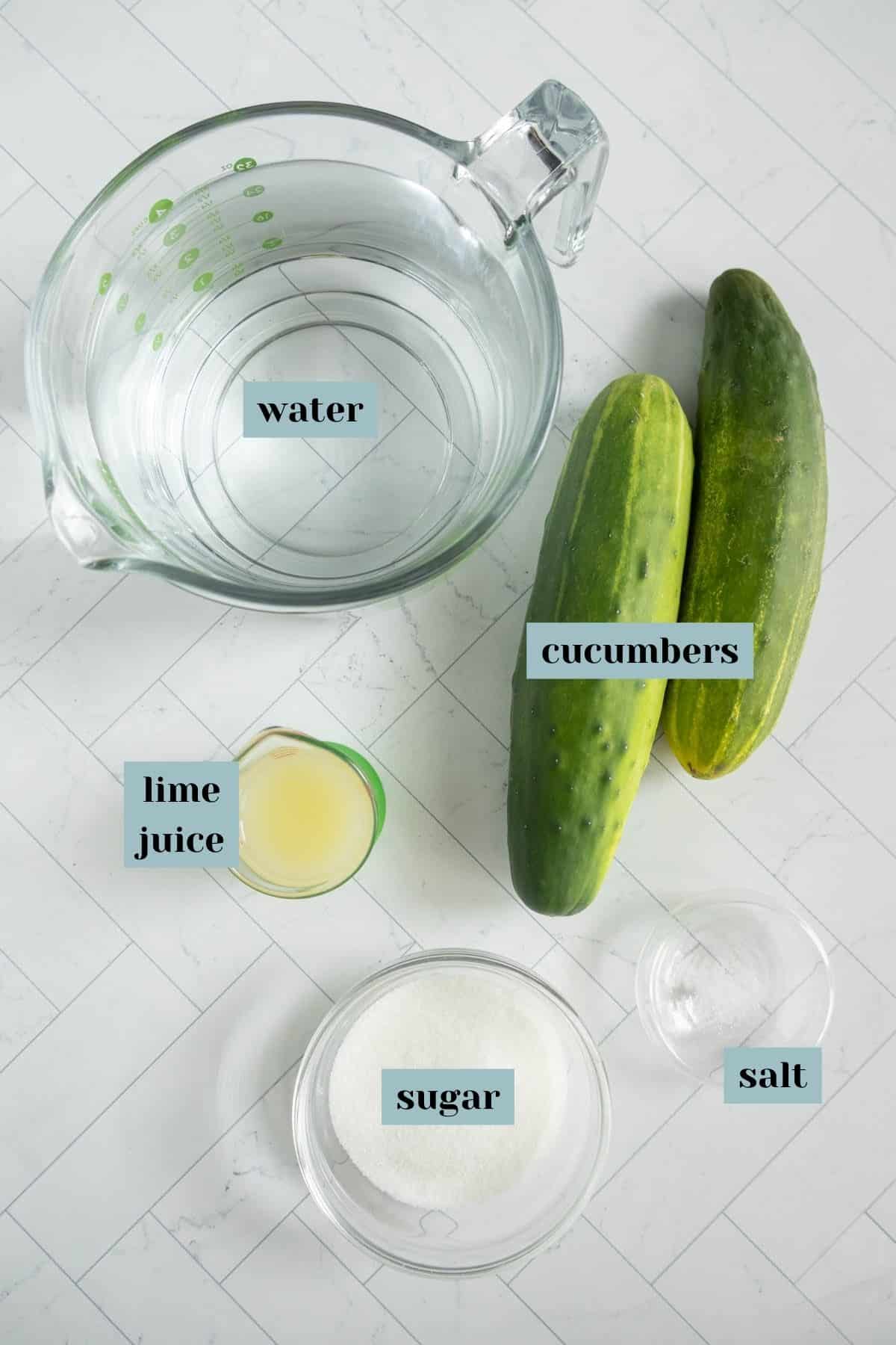 Ingredients for a recipe arranged on a countertop: a measuring cup with water, two cucumbers, a small container with lime juice, a bowl of sugar, and a small dish of salt.