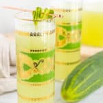 Two glasses of green-hued liquid garnished with cucumber slices, a cucumber beside them. The glasses feature intricate gold and green designs.