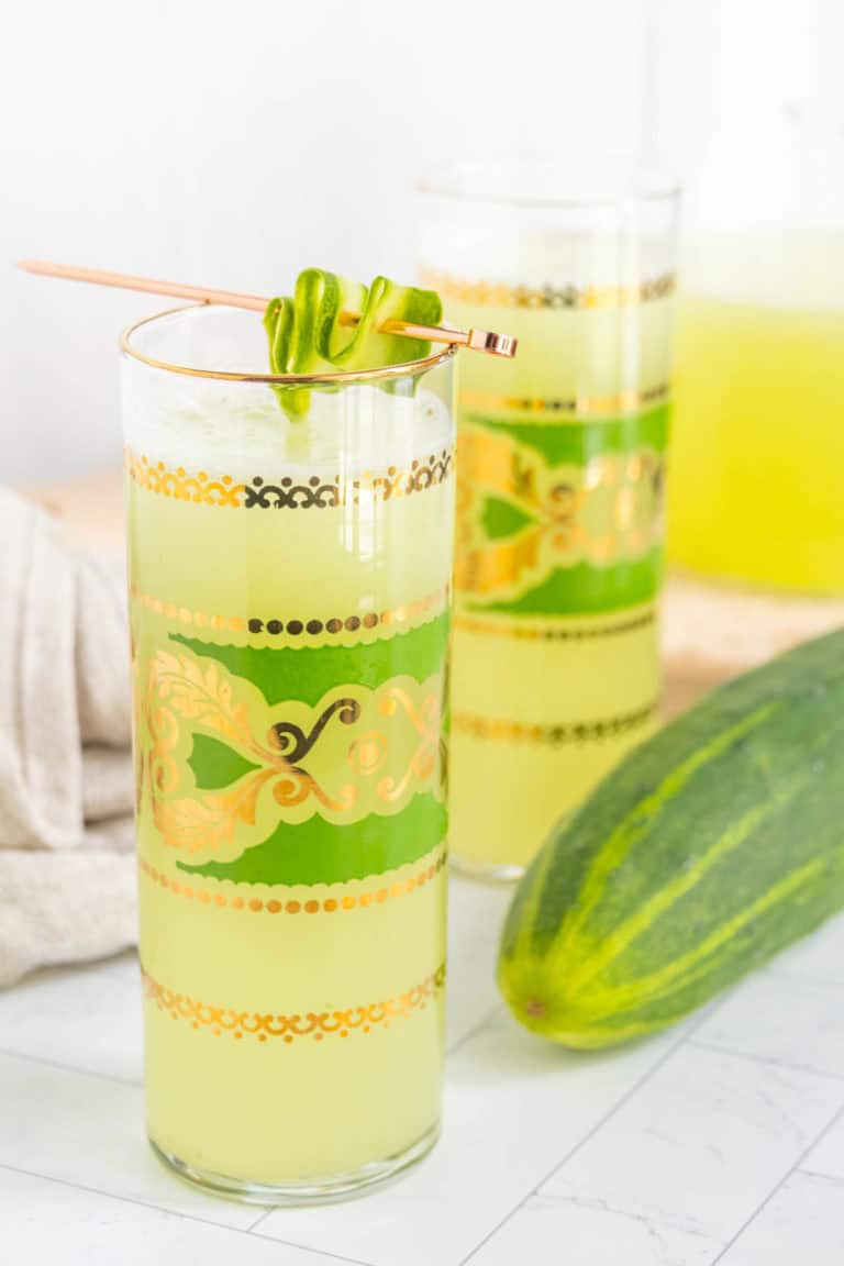 Two glasses of green-hued liquid garnished with cucumber slices, a cucumber beside them. The glasses feature intricate gold and green designs.