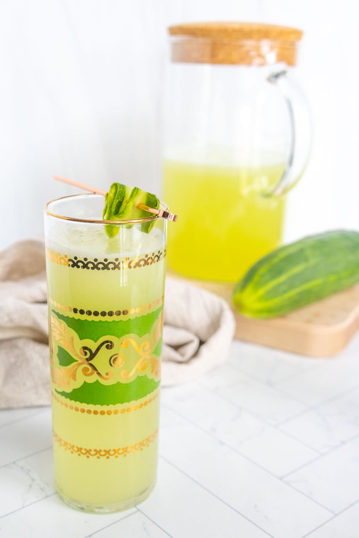 A glass of green juice garnished with cucumber slices on a toothpick, with a pitcher of the same juice and a whole cucumber in the background on a white countertop.