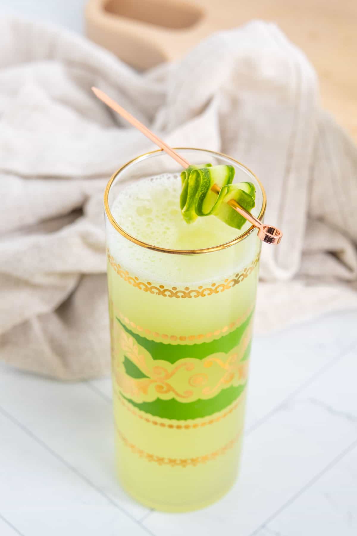 A tall glass filled with a green frothy beverage, garnished with two basil leaves on a rose gold stir stick, placed on a white surface with a beige cloth in the background.