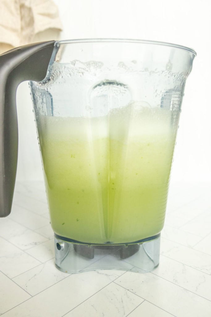 A blender pitcher filled with frothy, light green liquid sits on a tiled countertop.