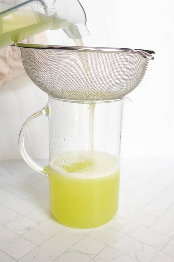 A light green liquid is being poured through a metal mesh strainer into a clear glass pitcher below.