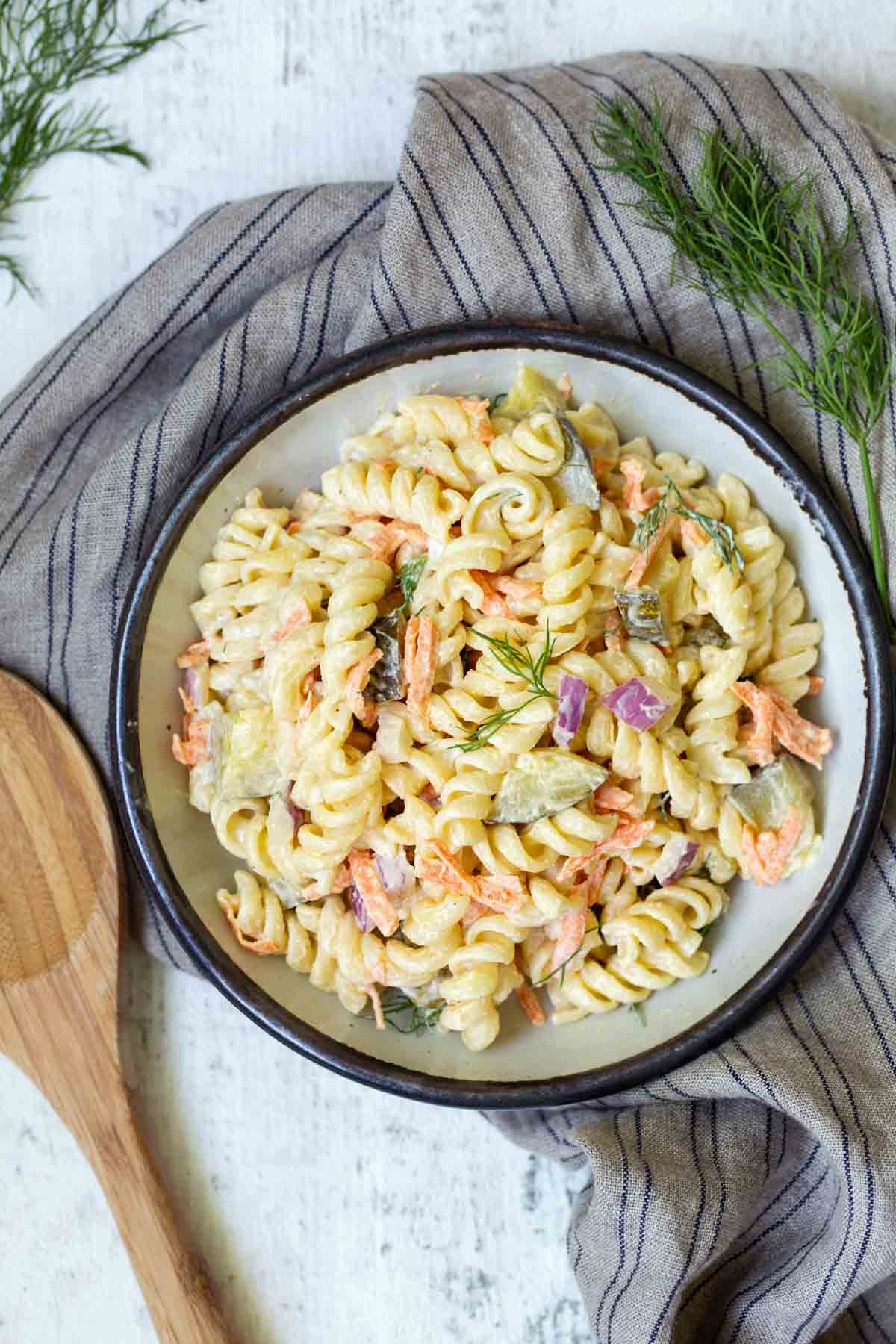 A bowl of pasta salad with rotini, diced vegetables, and a creamy dressing, garnished with fresh dill, is placed on a striped cloth with a wooden spoon beside it.