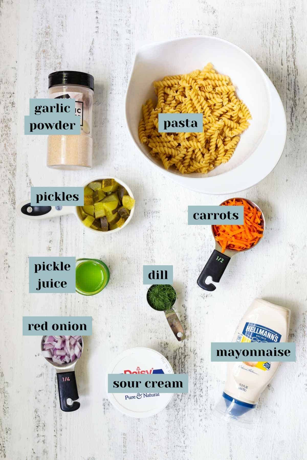 Overhead view of pasta salad ingredients including rotini pasta, garlic powder, pickles, carrot shreds, pickle juice, red onion, dill, sour cream, and mayonnaise on a white wood surface.