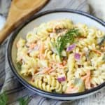 A bowl of creamy pasta salad with rotini, chopped vegetables, and garnished with a sprig of dill, placed on a cloth with a wooden spoon beside it.