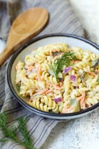 A bowl of creamy pasta salad with rotini, chopped vegetables, and garnished with a sprig of dill, placed on a cloth with a wooden spoon beside it.