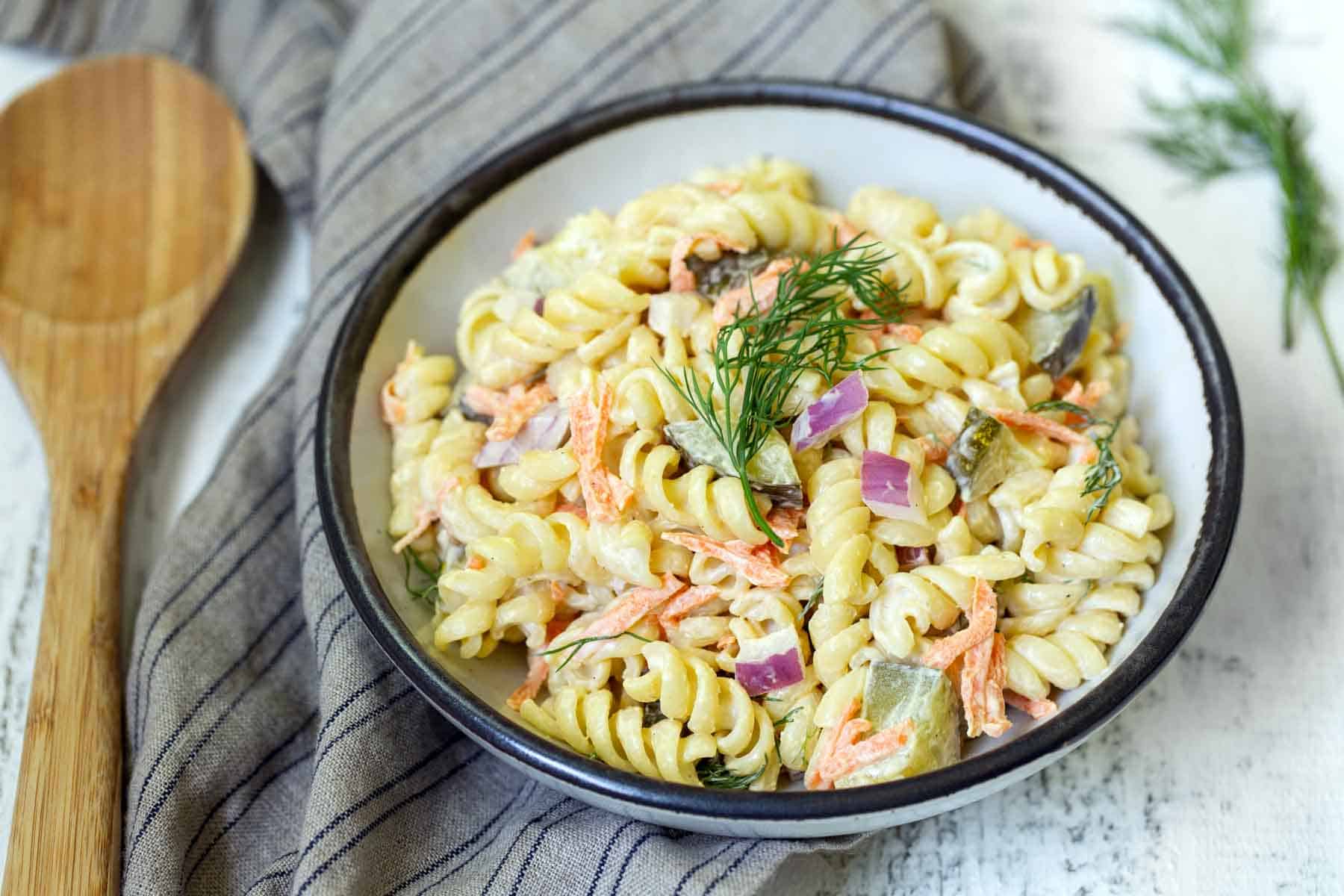 A bowl of creamy pasta salad with fusilli, shredded carrots, diced red onion, and fresh dill garnish, placed on a striped cloth with a wooden spoon beside it.