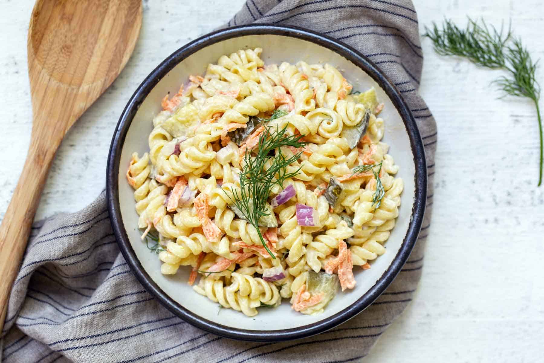 A bowl of rotini pasta salad with mixed vegetables and fresh dill garnish on a striped cloth, next to a wooden serving spoon.