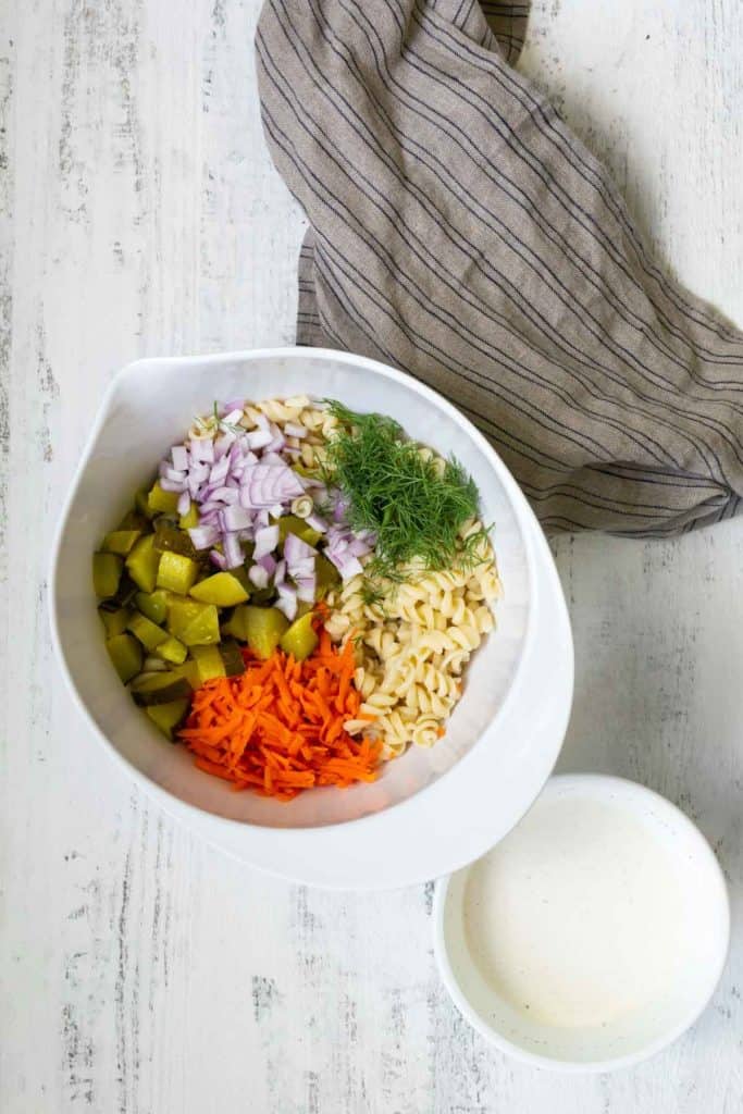 A white bowl contains chopped pickles, diced red onions, shredded carrots, pasta, and fresh dill. A small bowl of creamy dressing and a striped cloth napkin are placed nearby on a white surface.