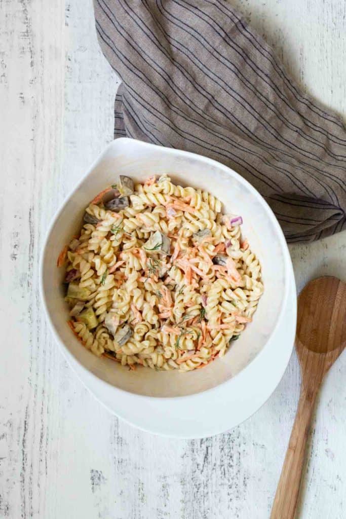 A bowl of rotini pasta salad with vegetables next to a wooden spoon and a striped cloth napkin on a white wooden table.