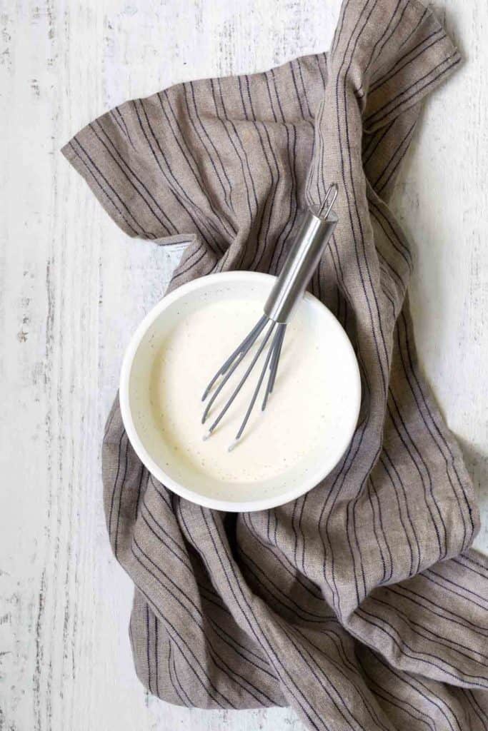 A whisk resting in a white bowl of cream on a striped cloth on a white wooden surface.