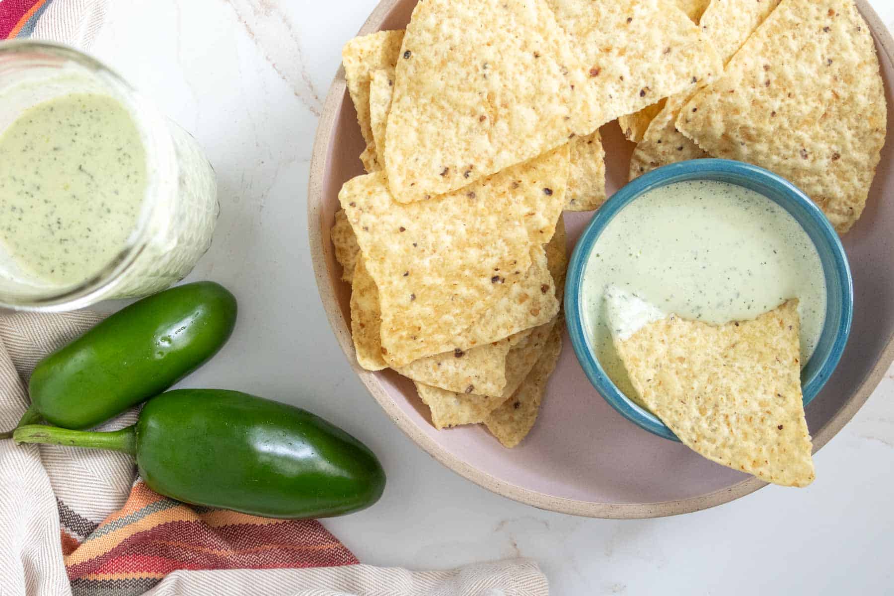 A plate of tortilla chips with one dipped in green sauce, next to a bowl of the same sauce, two fresh jalapenos, and a striped cloth.