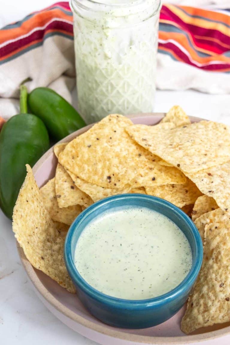 A bowl of tortilla chips next to a smaller bowl filled with a creamy green sauce. A striped cloth and two whole jalapeños are in the background, along with a jar containing more sauce.