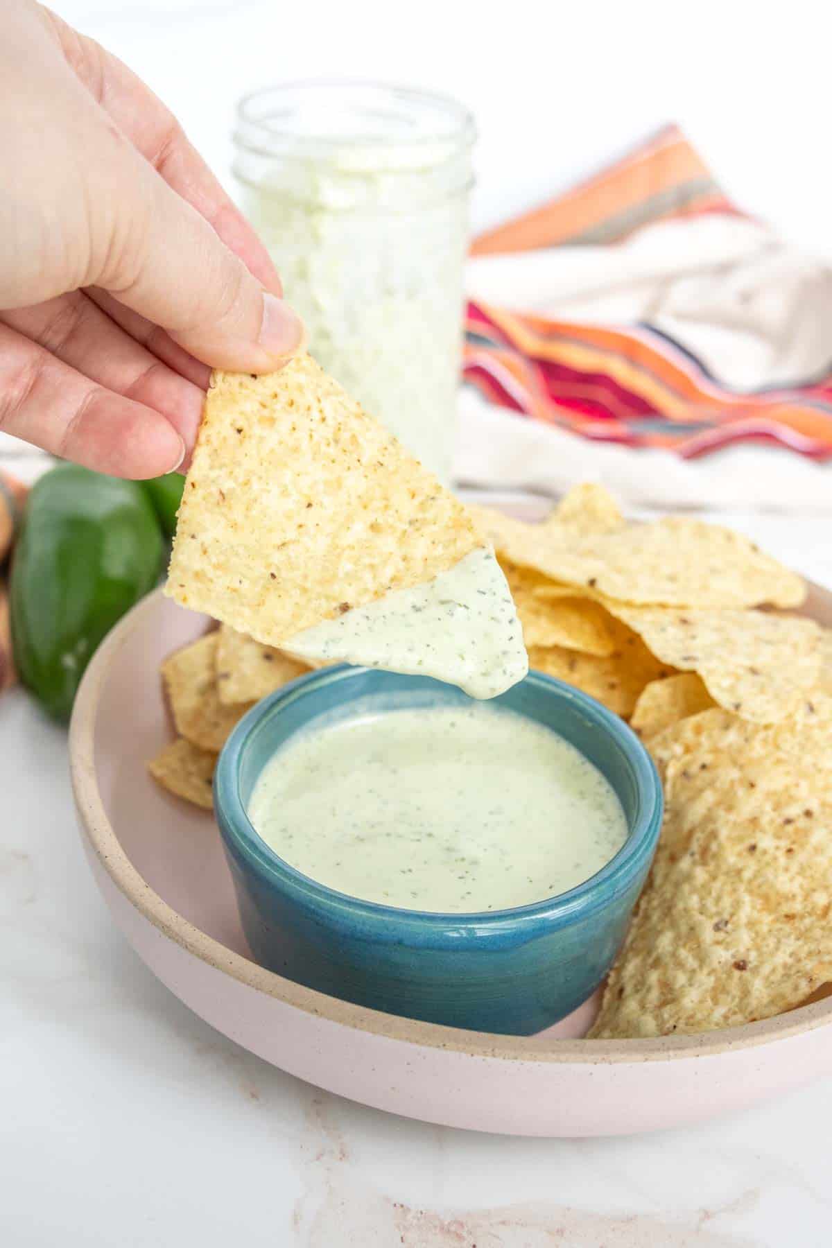 A hand dips a tortilla chip into a bowl of creamy dip. A plate of tortilla chips, a green pepper, and a jar with a green substance are in the background.