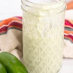 A jar filled with a creamy, white sauce with visible herbs. The jar is on a surface next to two green jalapenos and a striped cloth.